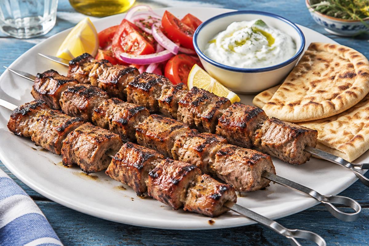 Skewers of charred pork cubes with tzatziki, pita, tomato, onion and lemon
